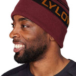 Flylow OG Pom Beanie 2024 - A maroon beanie with a brown pom-pom on top and an embroidered black band featuring orange lettering that reads "FLYLOW".