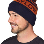 Flylow OG Pom Beanie 2024 - A navy blue beanie with an orange stripe and a brown pom-pom on top.