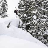 Lib Tech Skunk Ape Camber Snowboard 2025 - A snowboarder is riding a steep slope with trees in the background, wearing a black jacket and white pants.