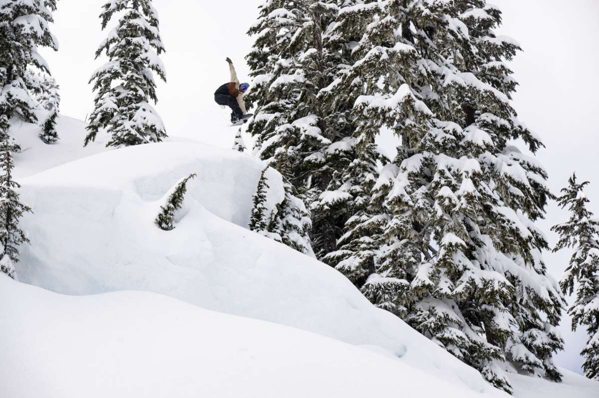Lib Tech Skunk Ape Camber Snowboard 2025 - A snowboarder is riding a steep slope with trees in the background, wearing a black jacket and white pants.