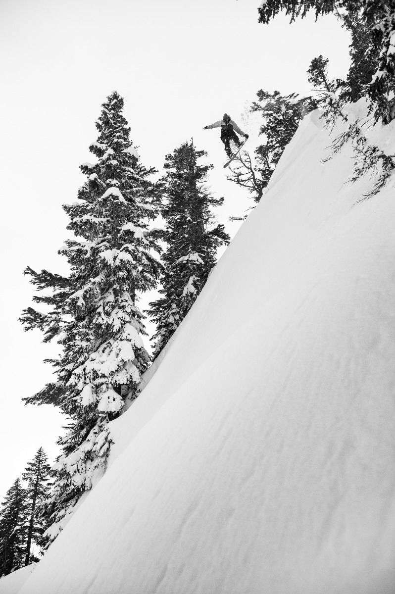 Lib Tech Skunk Ape Camber Snowboard 2025 - A snowboarder is seen from below, jumping off a steep snowy slope with trees in the background.