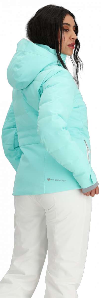 Obermeyer Women's Cosima Down Jacket 2024 - A woman with long dark hair wearing a light blue down jacket and white pants, facing left.