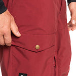 Quiksilver Utility Shell Pant 2023 - A pair of maroon snow pants with a black logo patch on the left leg featuring a white wave design and the word "Quiksilver" in white letters.