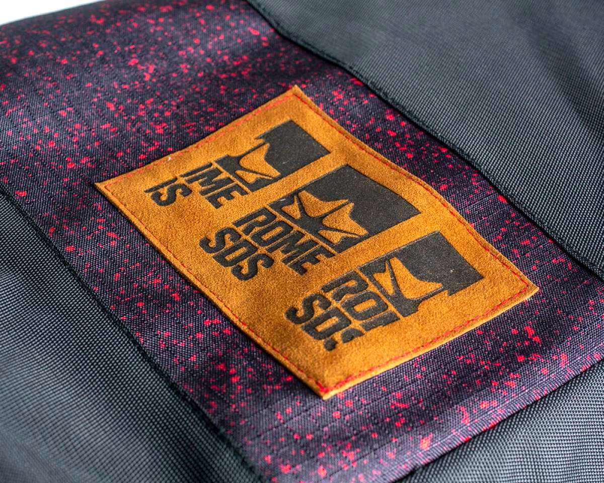 Rome Escort Wheelie Board Bag 2023 - A black bag with red speckles and a brown logo patch on the front.