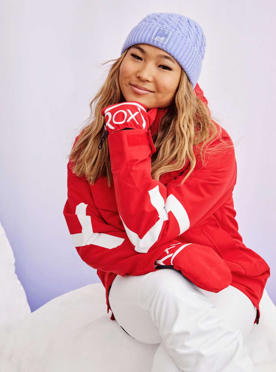 Roxy Women's Chloe Kim Overhead Jacket 2023 - A red jacket with a white logo on the sleeve and chest, worn by a woman posing in front of a snowy background.