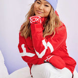 Roxy Women's Chloe Kim Overhead Jacket 2023 - A red jacket with a white logo on the sleeve and chest, worn by a woman posing in front of a snowy background.