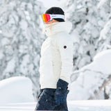 Roxy Women's Dusk Warmland Insulated Jacket 2024 - A white jacket with a black logo on the left sleeve and a hood, worn by a woman standing in front of snow-covered trees.