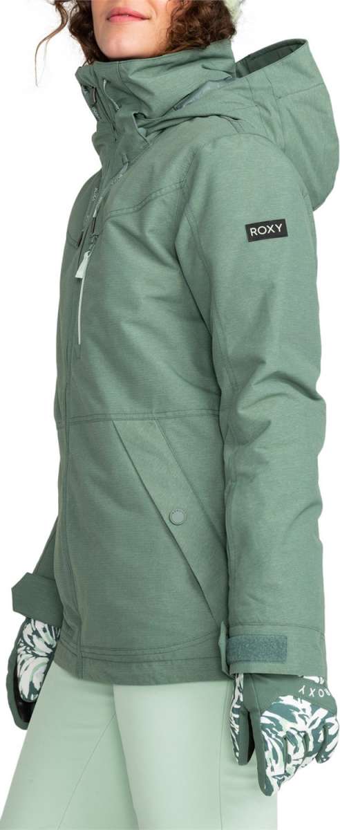 Roxy Women's Presence Insulated Parka Jacket 2024 - A woman with curly brown hair is wearing a light green parka jacket with a hood and a black rectangular patch on the left sleeve that says "ROXY" in white letters.