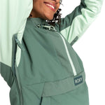Roxy Women's Radiant Lines Pullover Jacket 2024 - A woman with curly dark hair wearing a green jacket with white stripes down the front and a drawstring hood, posing in front of a light gray background.