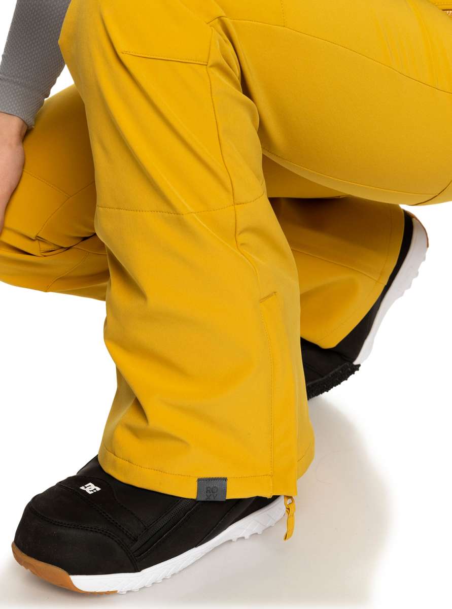 Roxy Women's Rising High Pant 2023 - A pair of yellow snow pants with a zippered pocket on the side and a small black tag near the hem.