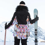 Roxy Women's Stated Insulated Jacket 2023 - A black jacket with a colorful abstract floral pattern on the back and sleeves, worn by a woman standing in front of snowy mountains.