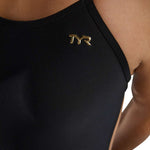 TYR Women's Cutoutfit Torch Swimsuit - A black swimsuit with a cut-out design on the sides, featuring a gold logo that says "TYR" in bold letters.