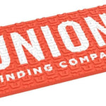 Union Surf Stomp Pad - A coral-colored pad with a textured surface, featuring a repeating pattern of stylized "U" shapes and the word "UNION" in white letters at the top.