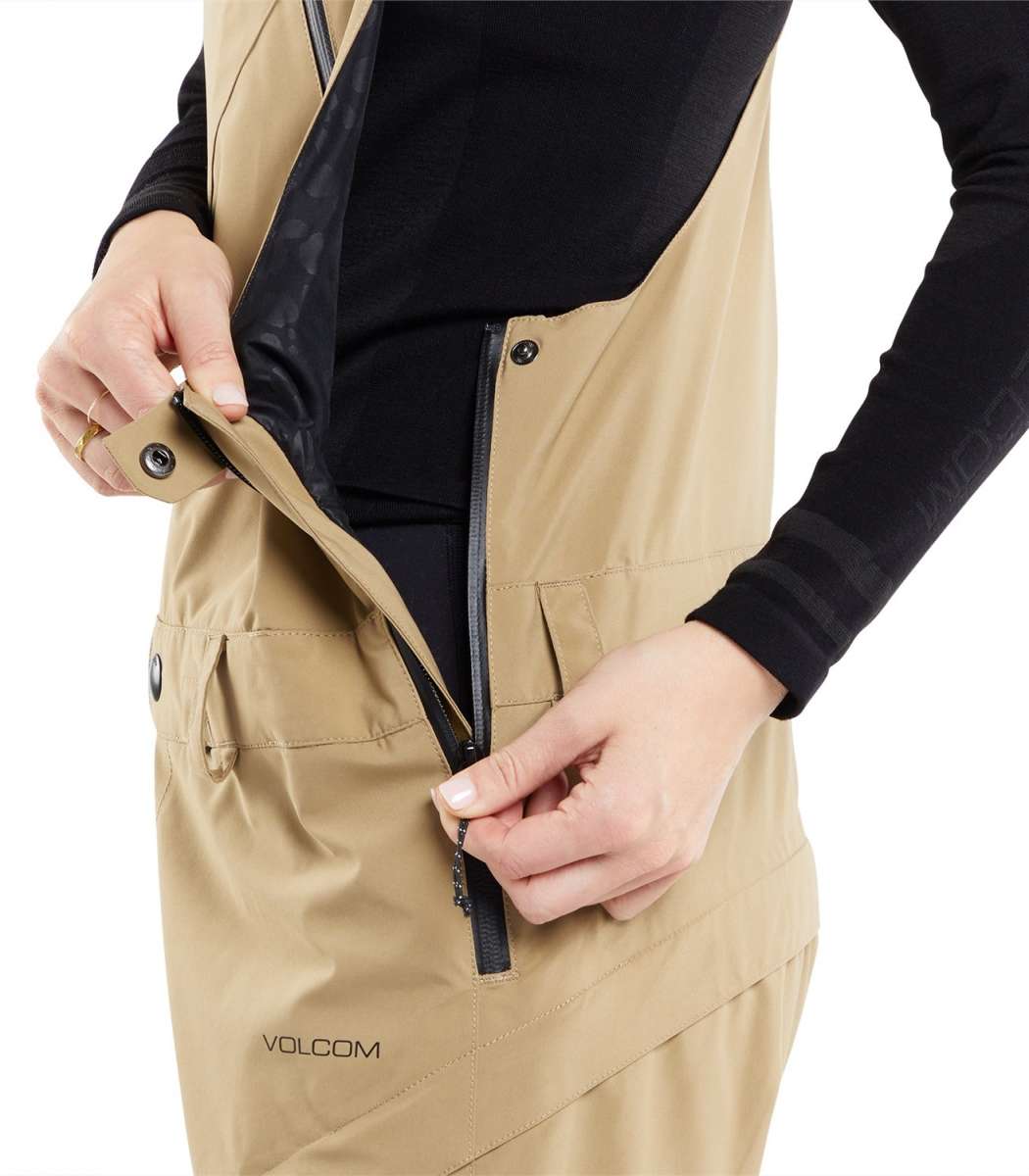 Volcom Women's Elm Stretch GORE-TEX Bib Pant - A tan bib pant with a black zipper and snap buttons, featuring a diagonal pattern of darker tan lines on the side pockets.