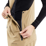 Volcom Women's Elm Stretch GORE-TEX Bib Pant - A tan bib pant with a black zipper and snap buttons, featuring a diagonal pattern of darker tan lines on the side pockets.