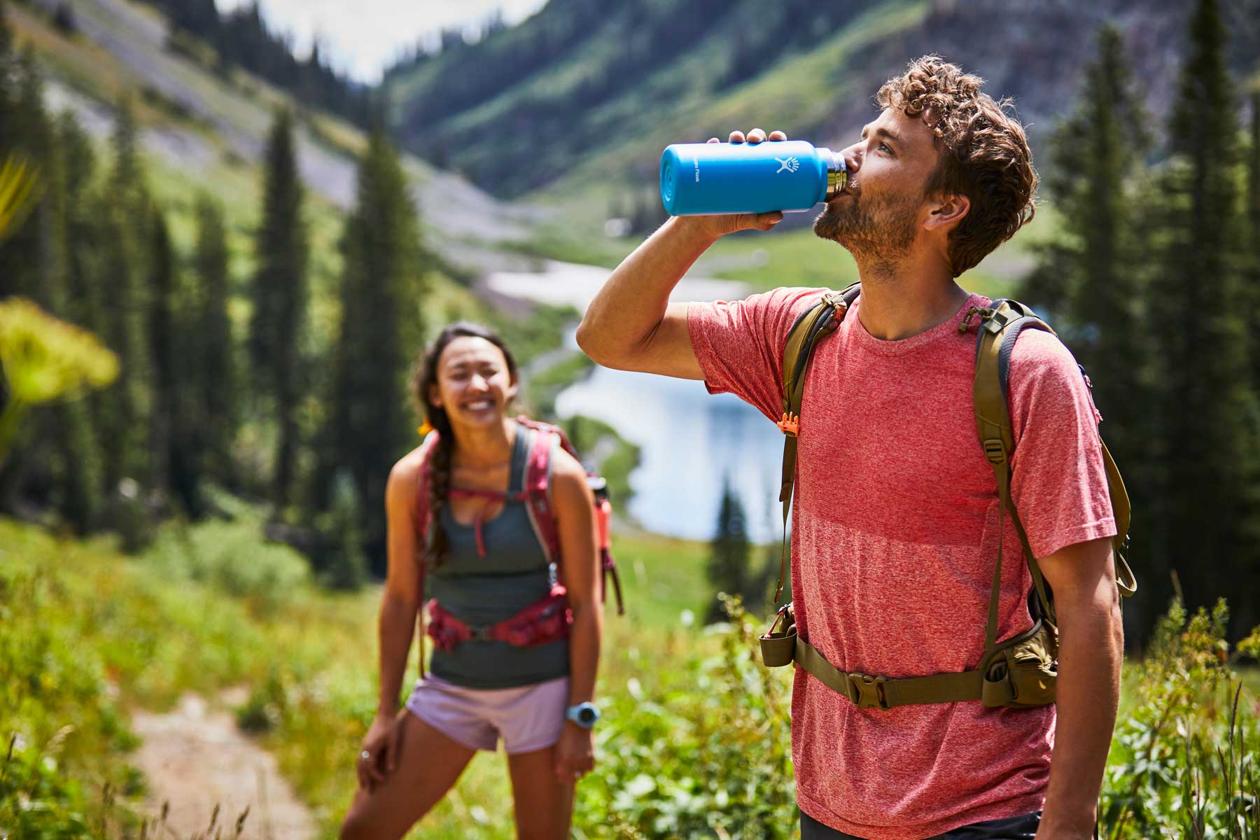 Hydro Flask 32oz Wide Mouth Water Bottle - A blue water bottle with a white lid, held by a man in a red shirt and green backpack strap over his right shoulder.