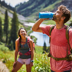 Hydro Flask 40oz Wide Mouth Water Bottle - A blue water bottle with a white lid, held by a man in a red shirt and green backpack strap over his right shoulder.